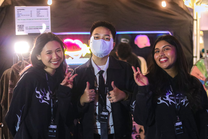 Three Club Cosmo's team members posing together with peace signs, one wearing a face mask, in front of a colorful background at an 88rising night market event.