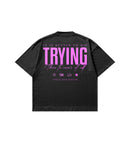 A black short-sleeve t-shirt shown from the back, featuring a large graphic print in pink text that reads 'IT IS BETTER TO DIE TRYING than to never at all.' with Club Cosmos icons below.
