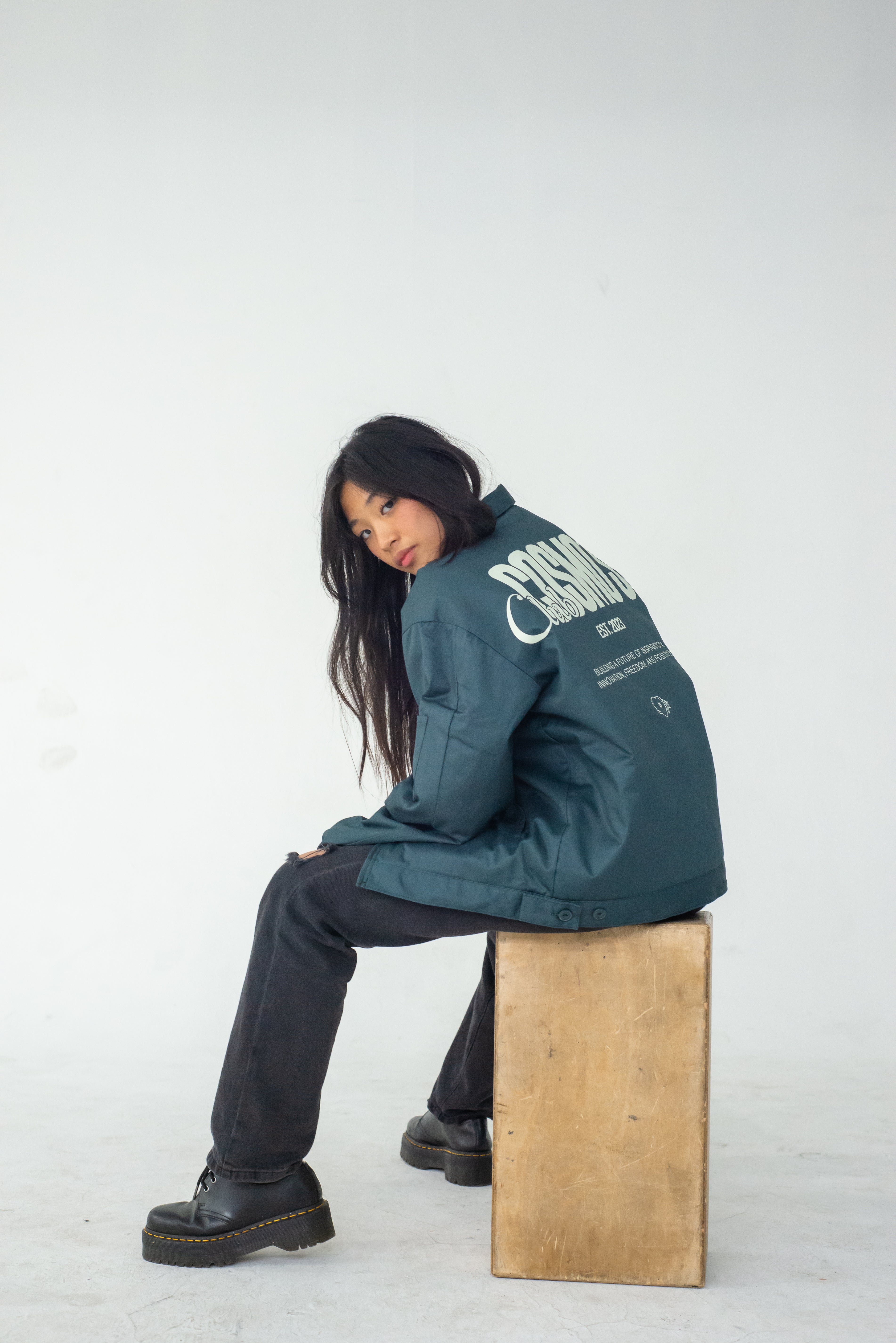 Model wearing a green Club Cosmo's utility jacket with text on the back, sitting on a wooden block against a white background.