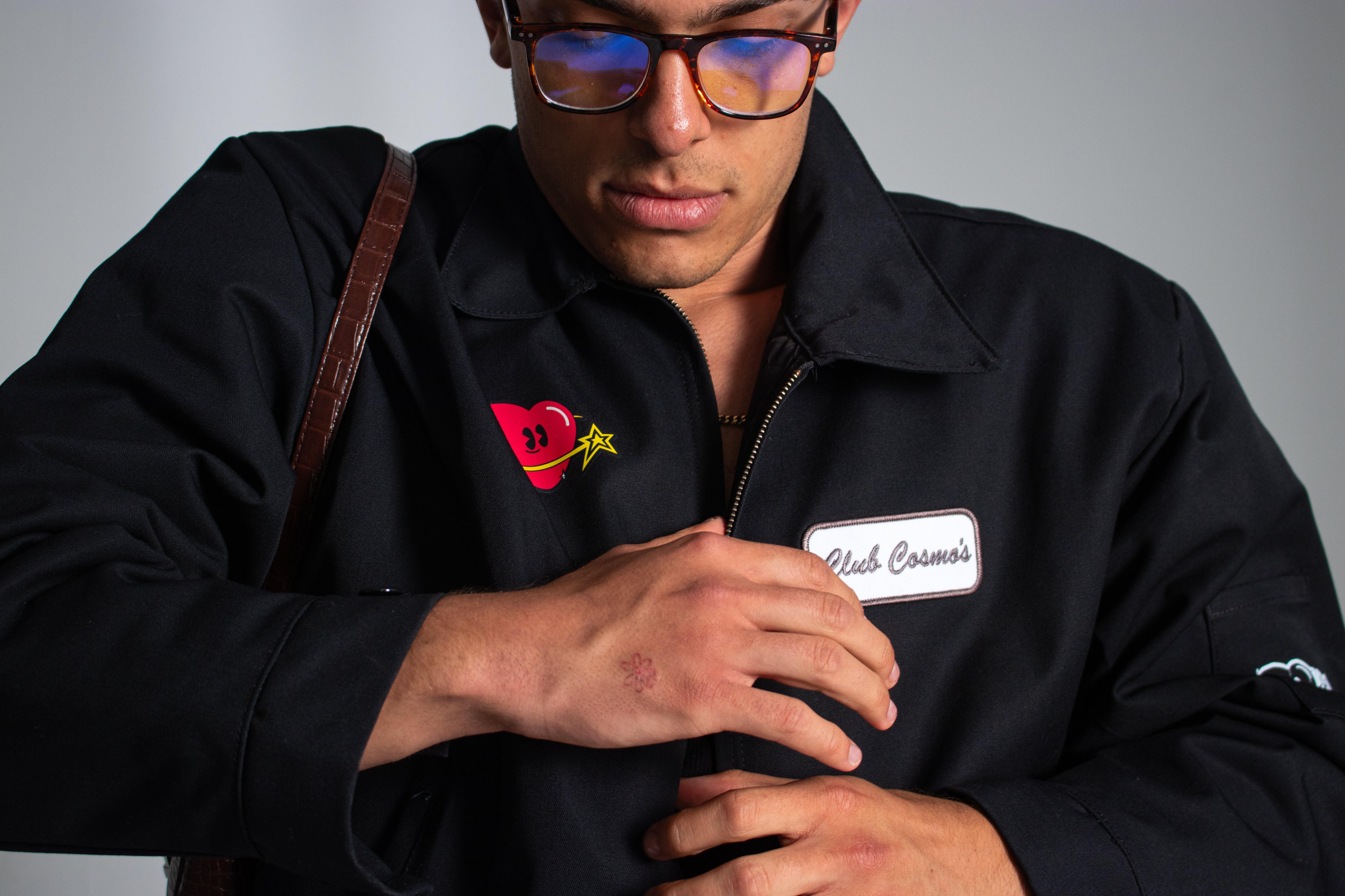 Model wearing a black Club Cosmo's utility jacket with a heart and star logo and embroidered patch on a white background.
