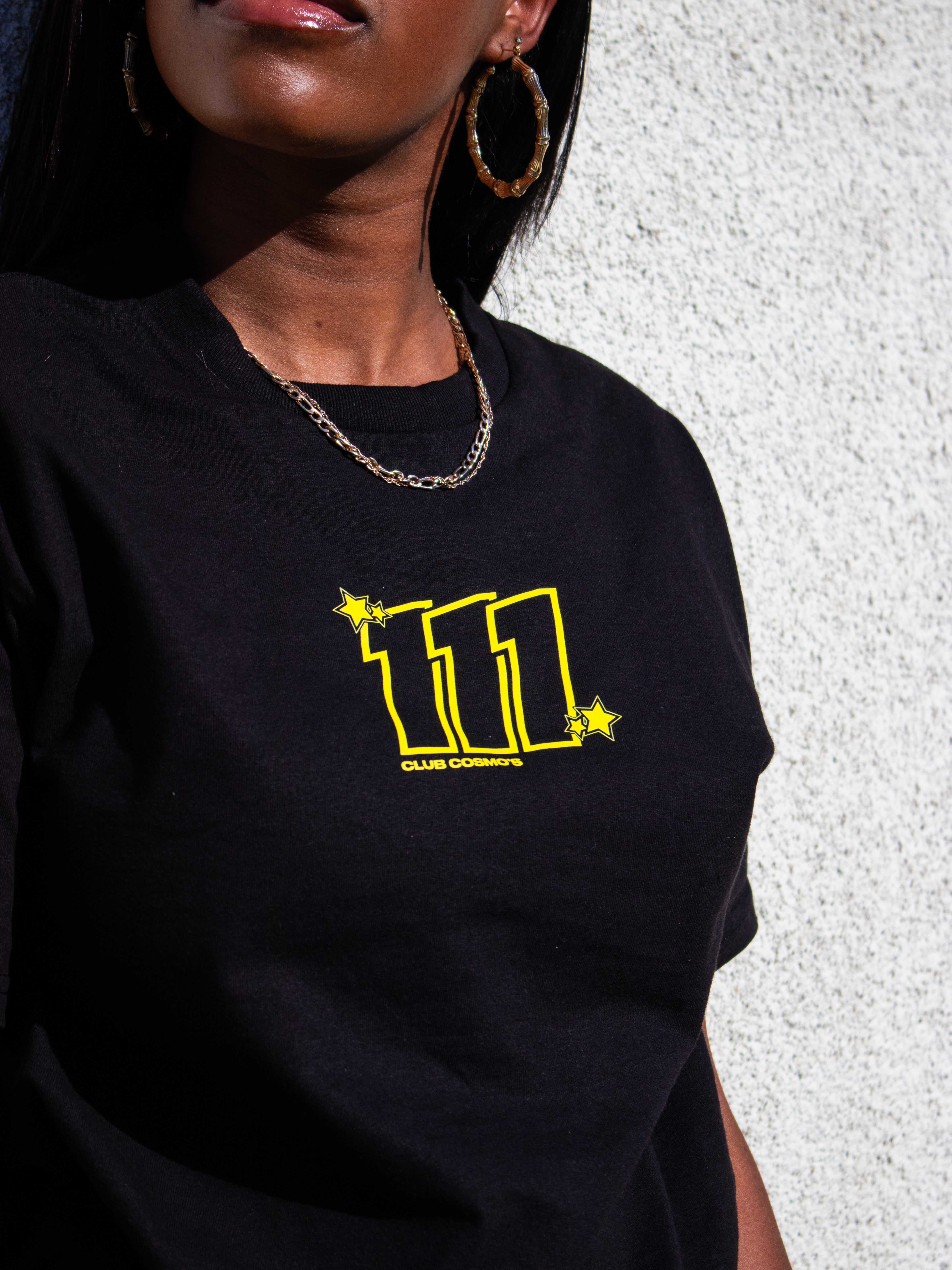 Model wearing a black t-shirt with yellow text '111 Club Cosmo's' against a gray background
