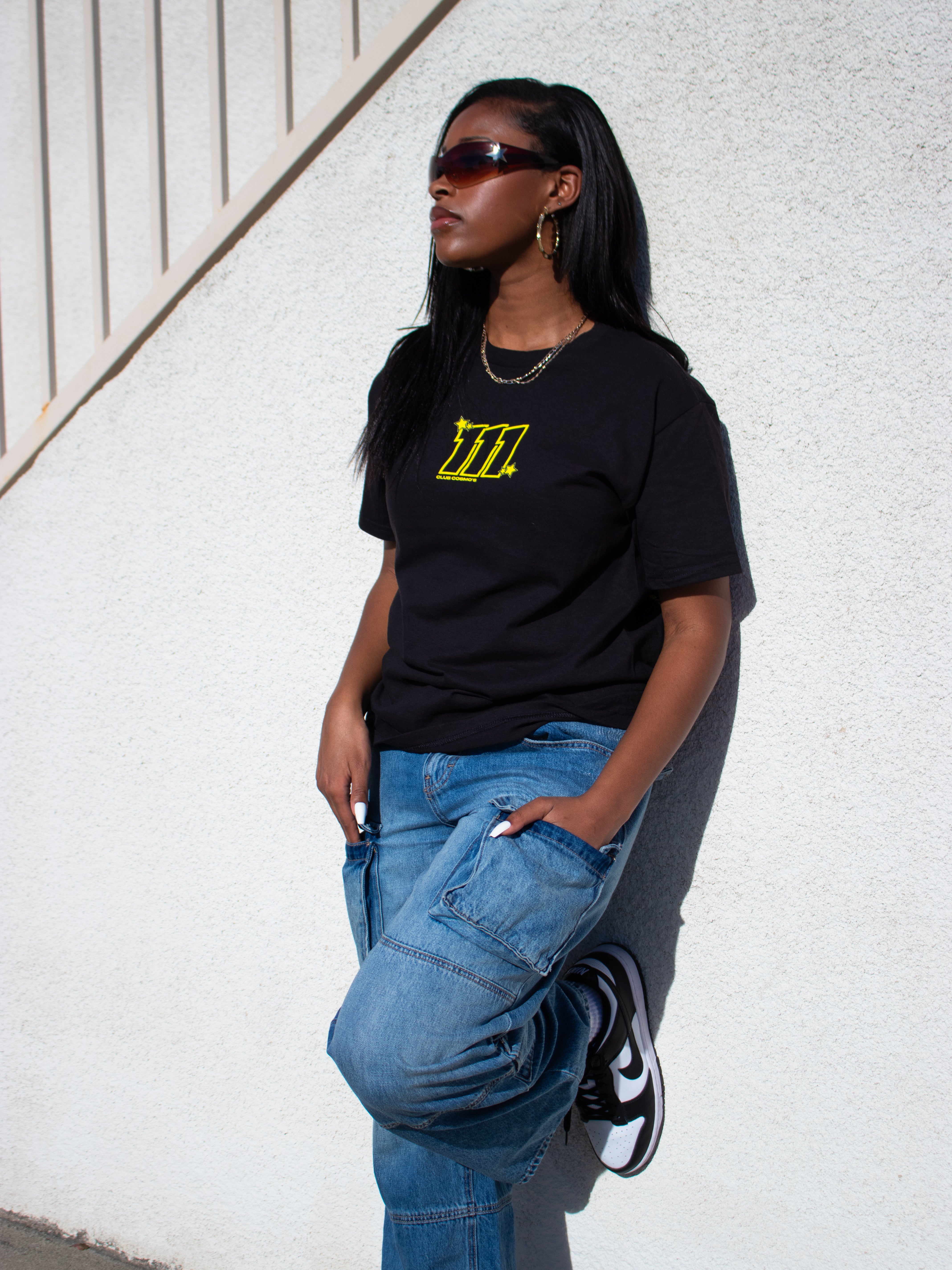 Model wearing a black t-shirt with a yellow '111 Club Cosmo's' logo and blue jeans against a white wall.