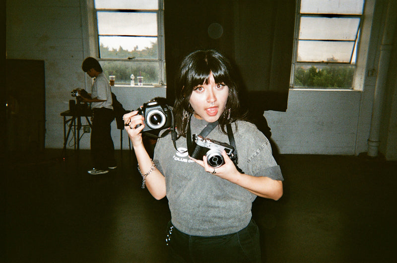 Club Cosmo's b roll specialist wearing a washed tee holding two cameras in a studio setting with large windows.