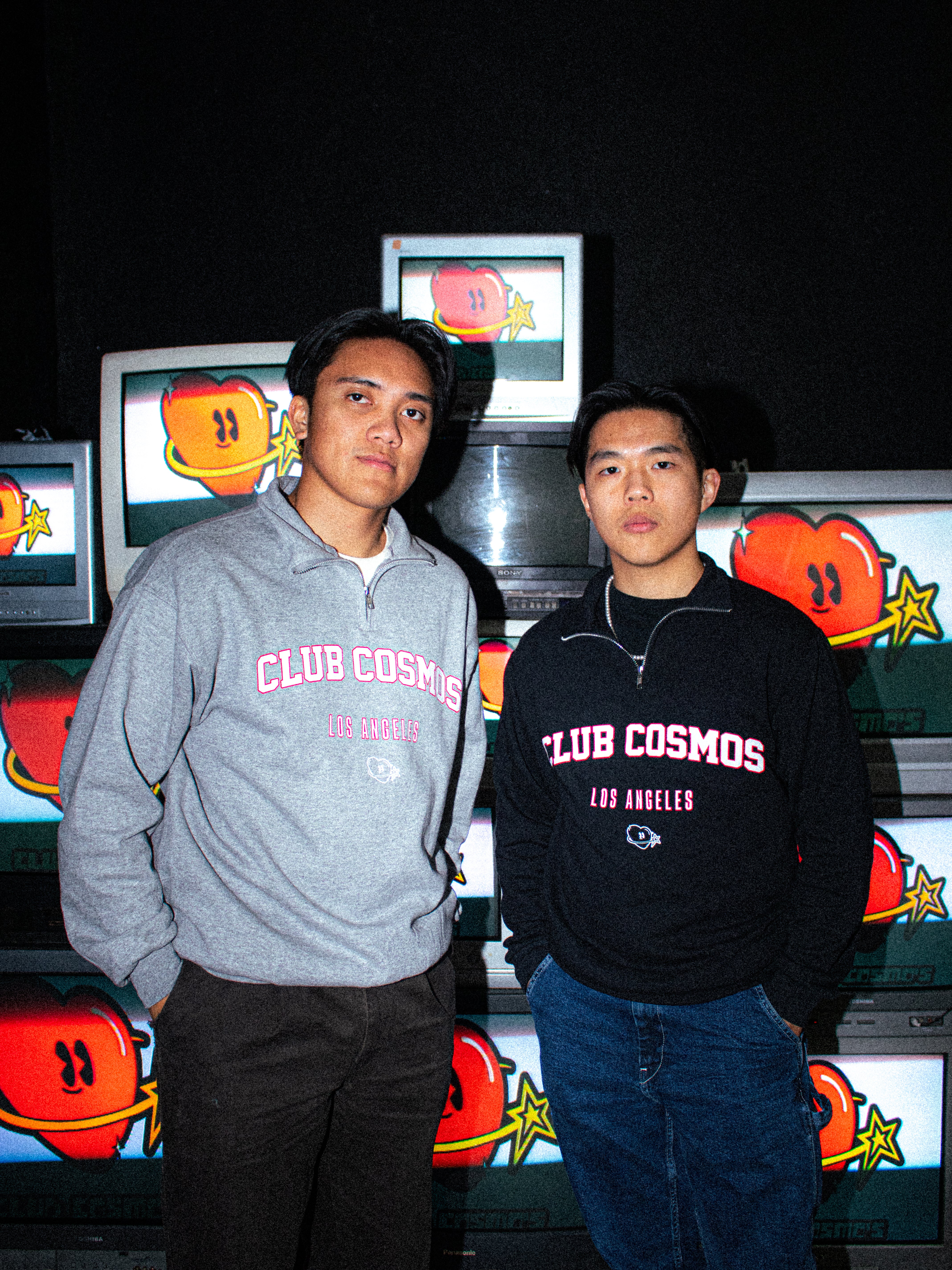 Two individuals wearing Club Cosmos quarter zip sweatshirts standing in front of vintage television screens.