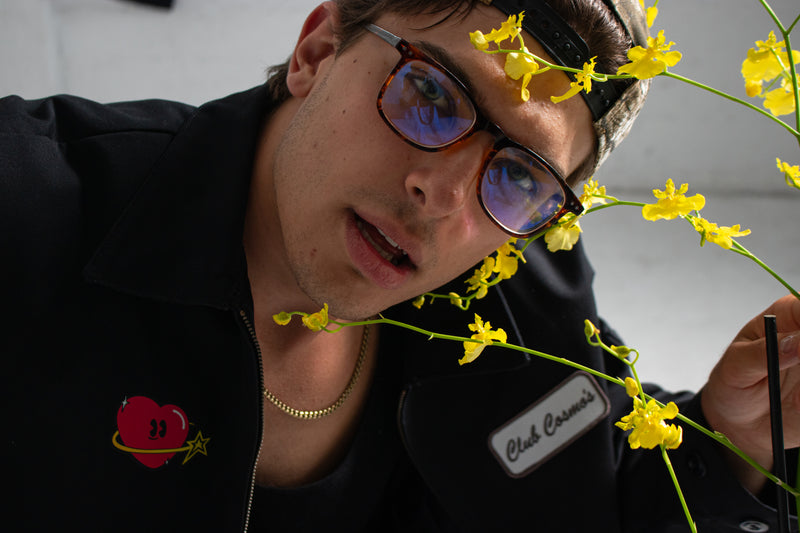 Model wearing glasses and a black Club Cosmo's utility jacket with yellow flowers in the foreground.