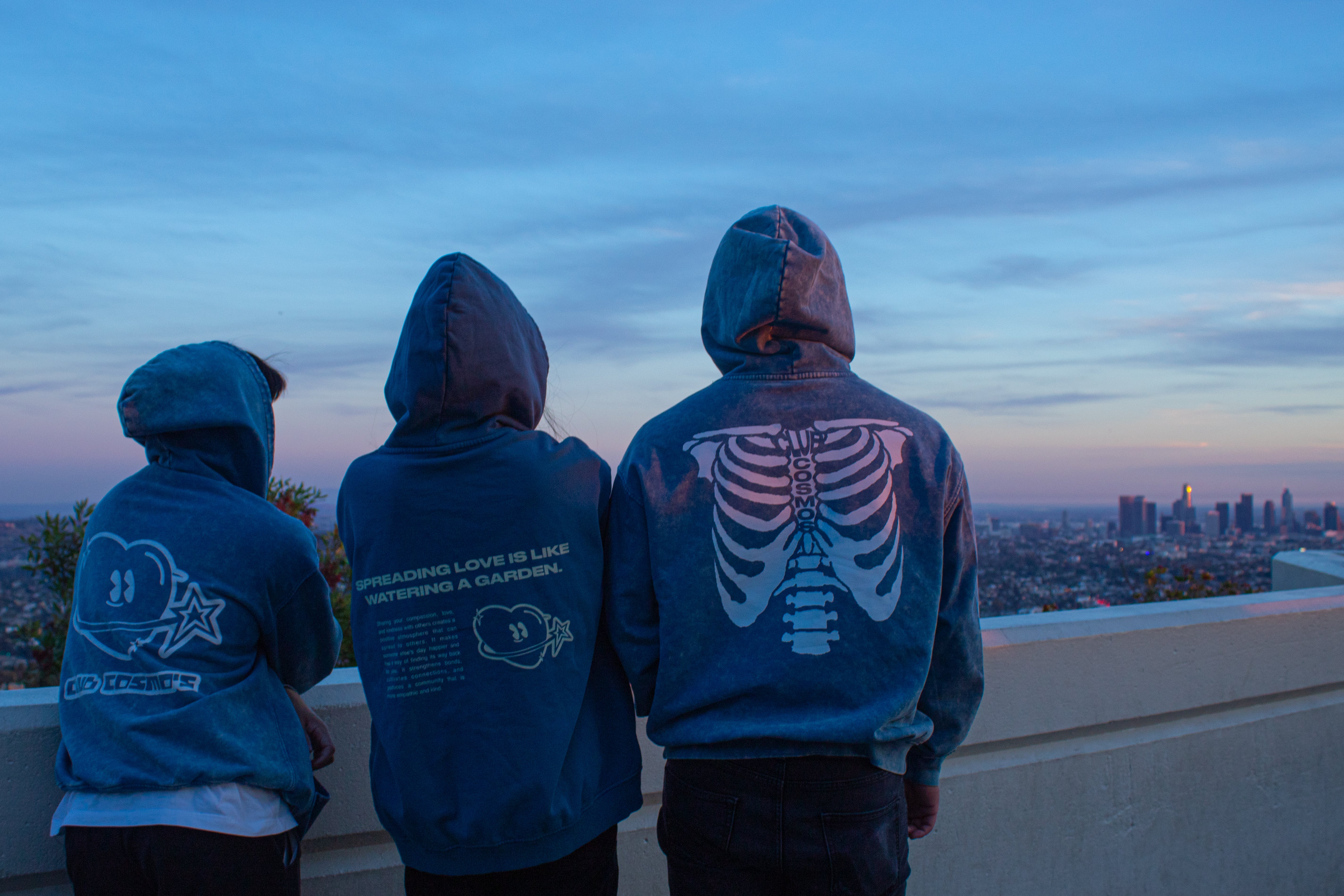 Three people wearing blue Club Cosmo's hoodies with graphic designs, standing on a rooftop overlooking a cityscape at sunset.
