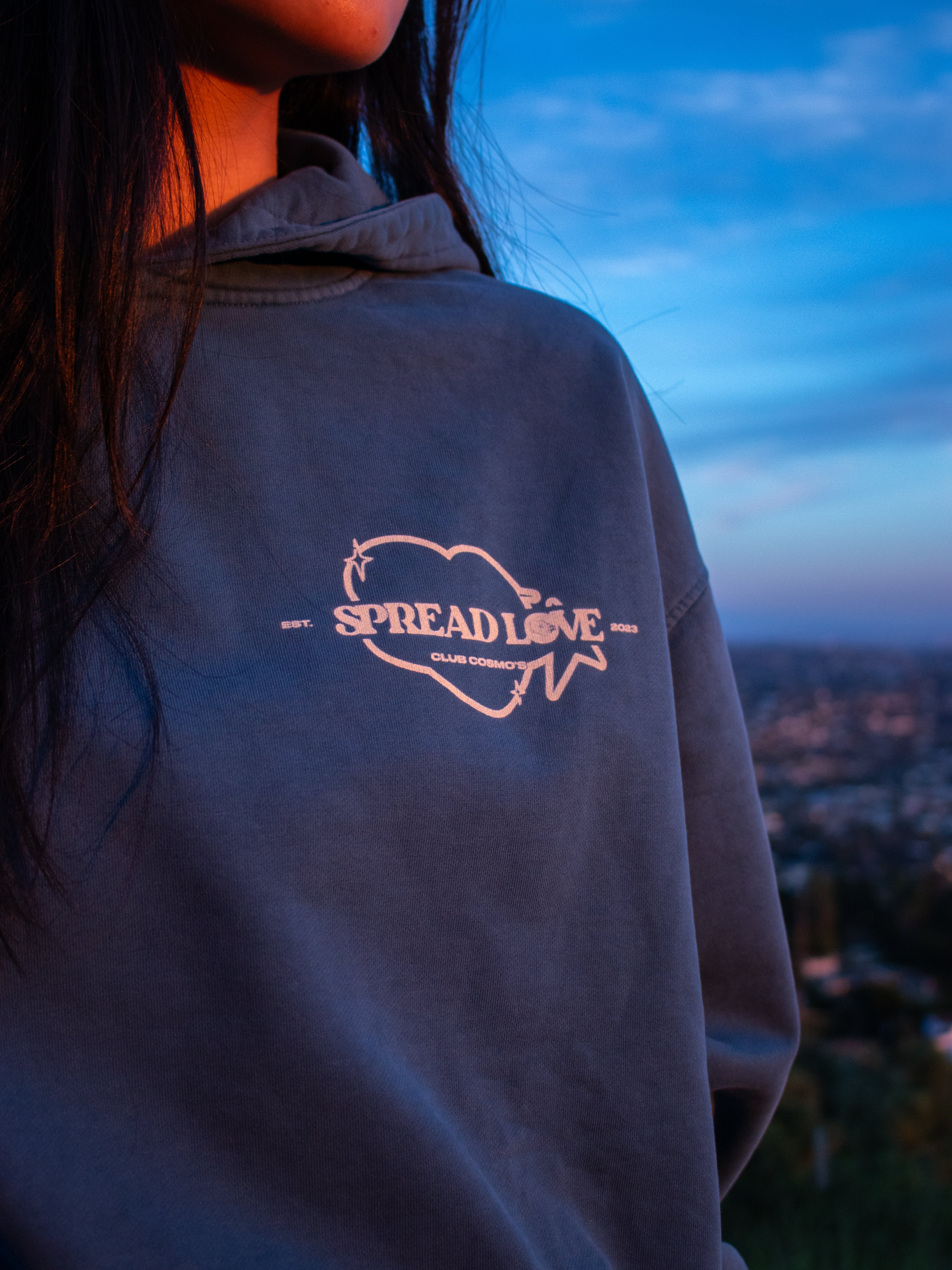 Model wearing a blue hoodie with 'Spread Love' and Club Cosmo's logo against a sunset sky.