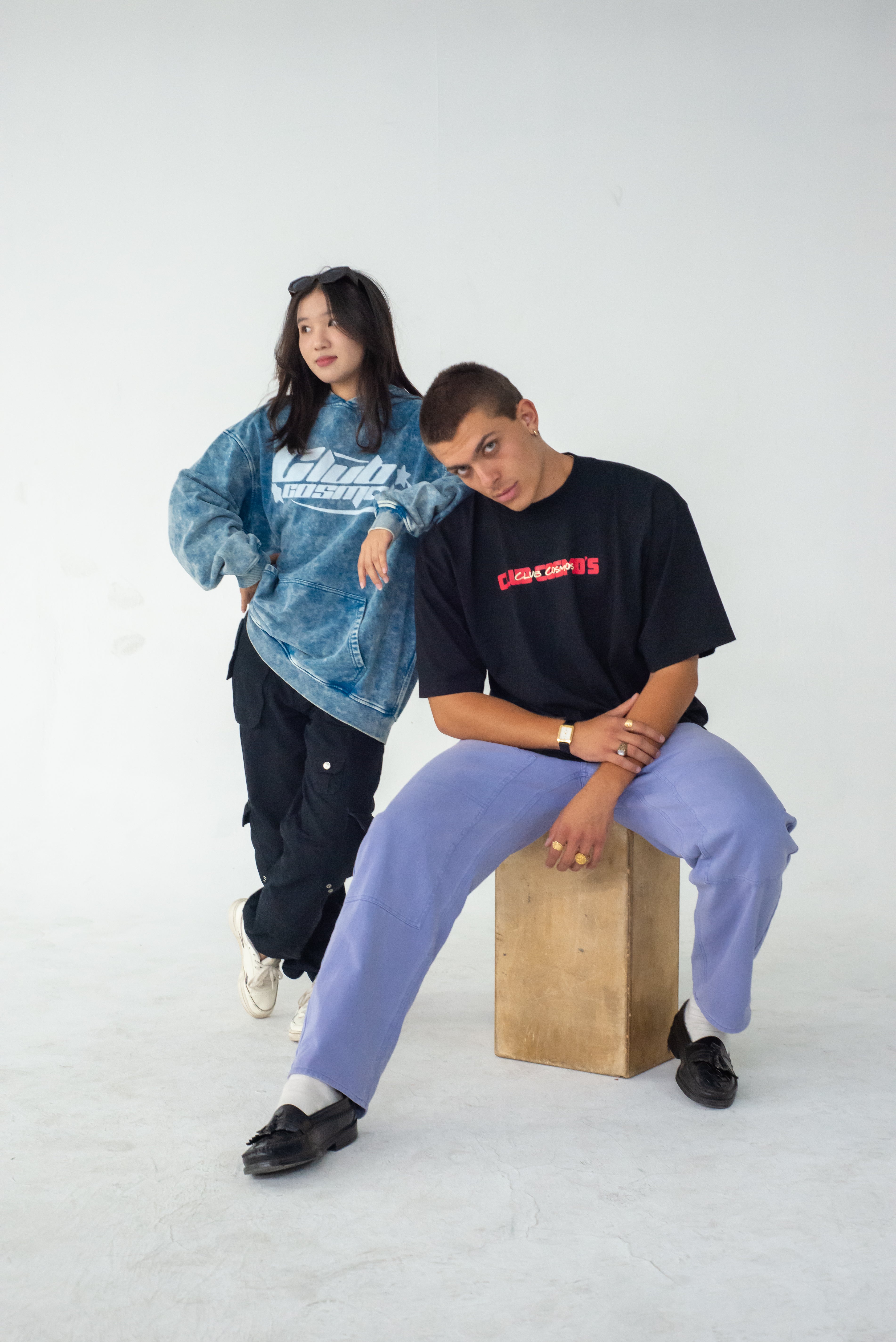 Two people posing, one with a blue Club Cosmo's hoodie and another with a black tee with Club Cosmos branding against a white background, one sitting on a wooden block.