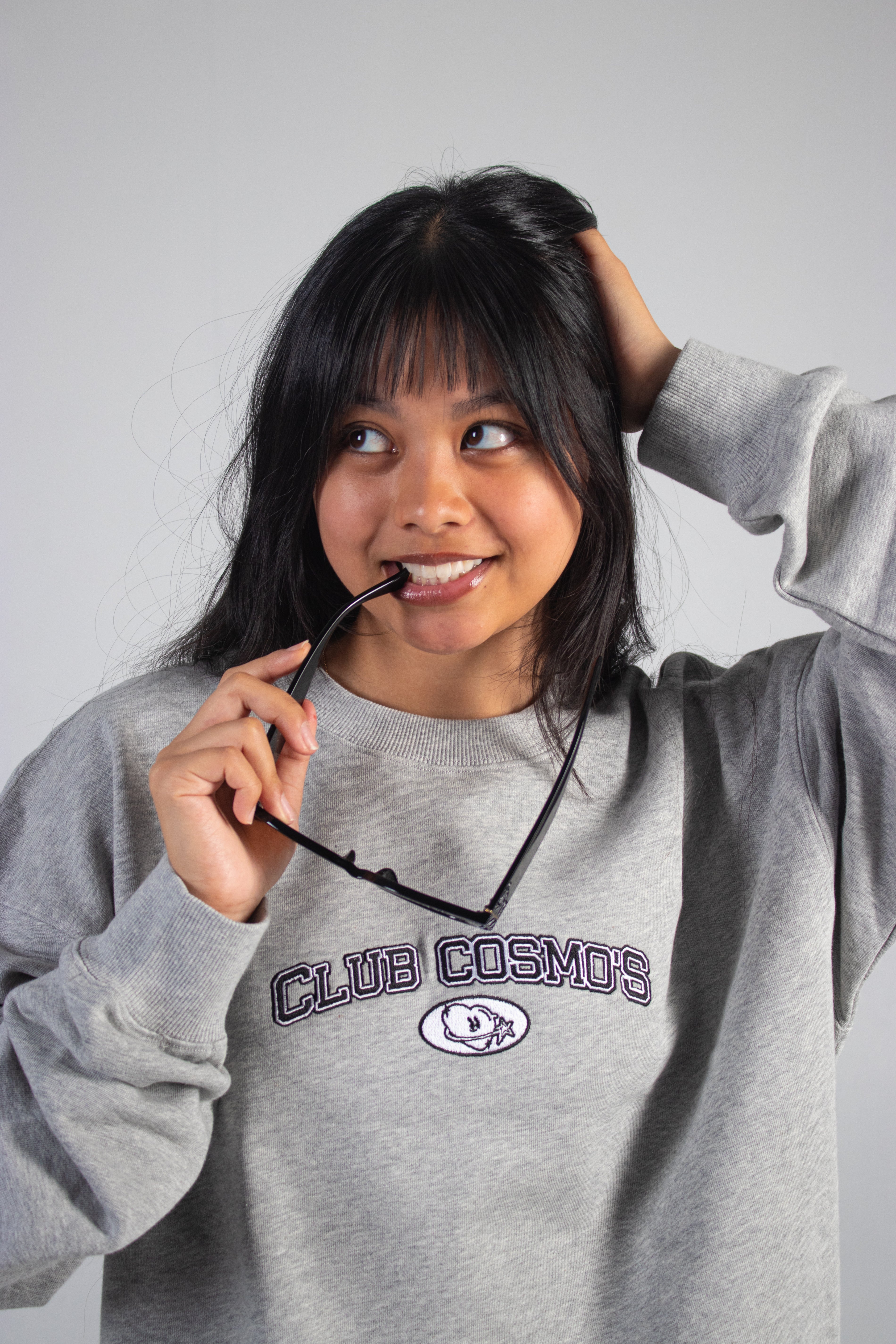 Model wearing a grey sweatshirt with 'CLUB COSMO'S' embroidered text and logo, holding sunglasses.