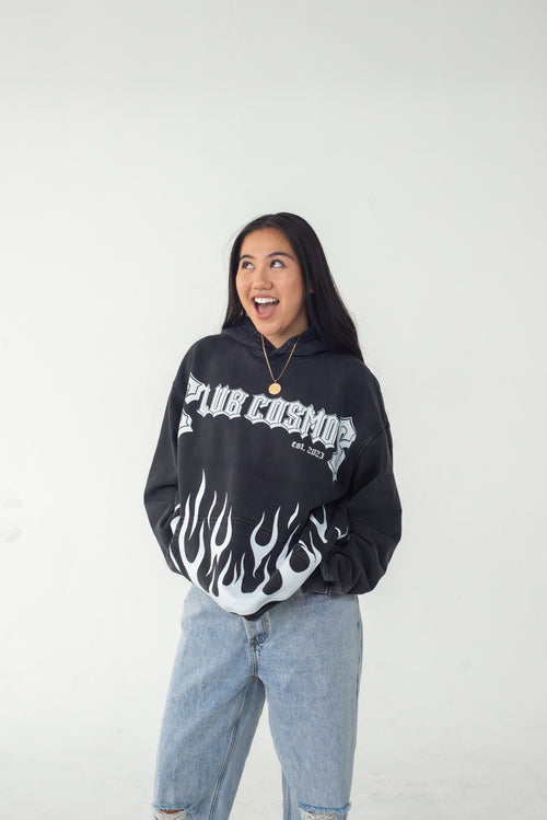 Model wearing a black hoodie with white text 'Club Cosmo's' and flames, standing against a white background.