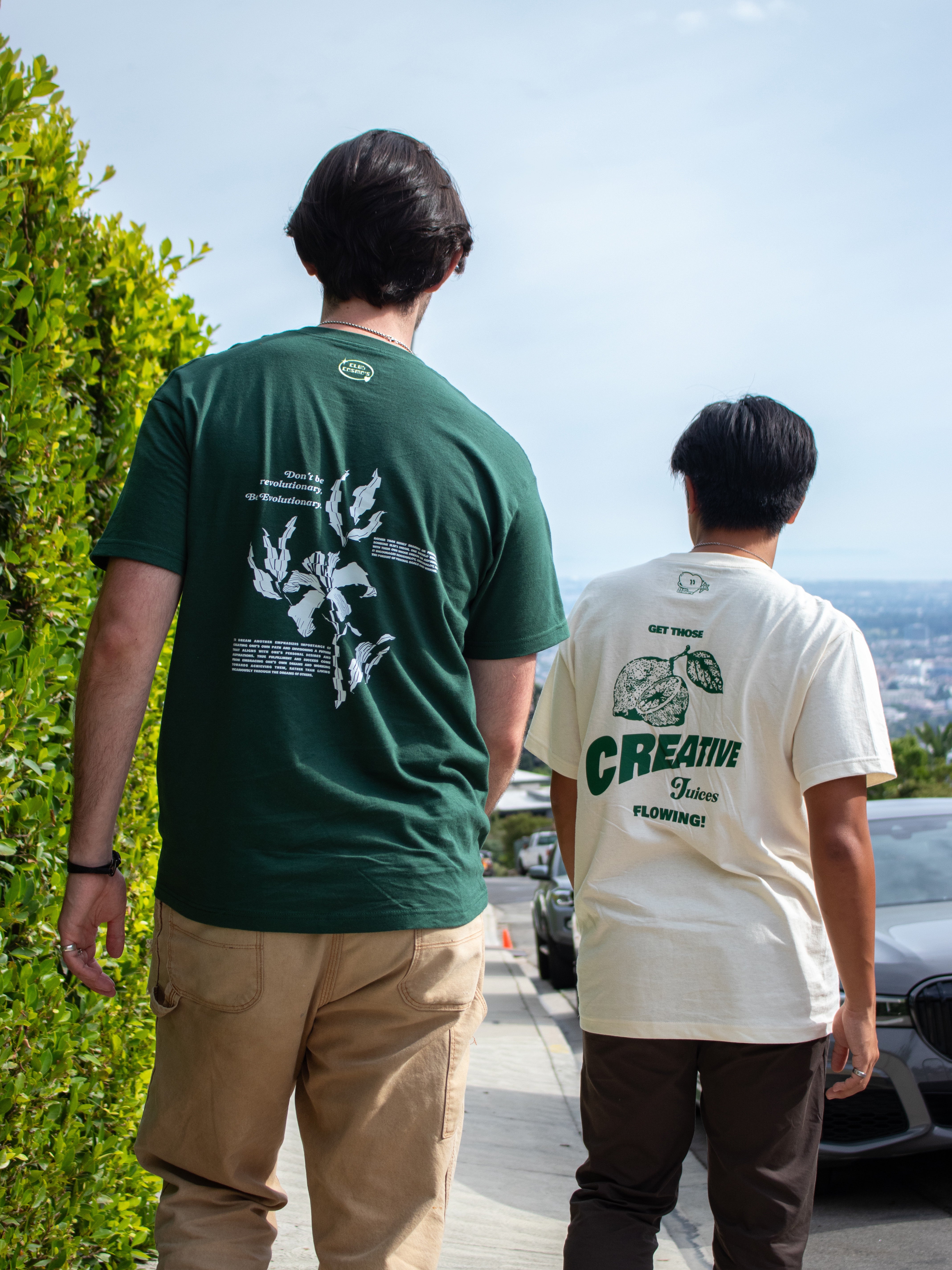 Two people wearing green and beige t-shirts with Club Cosmo's designs and text, standing outdoors overlooking the city skyline.