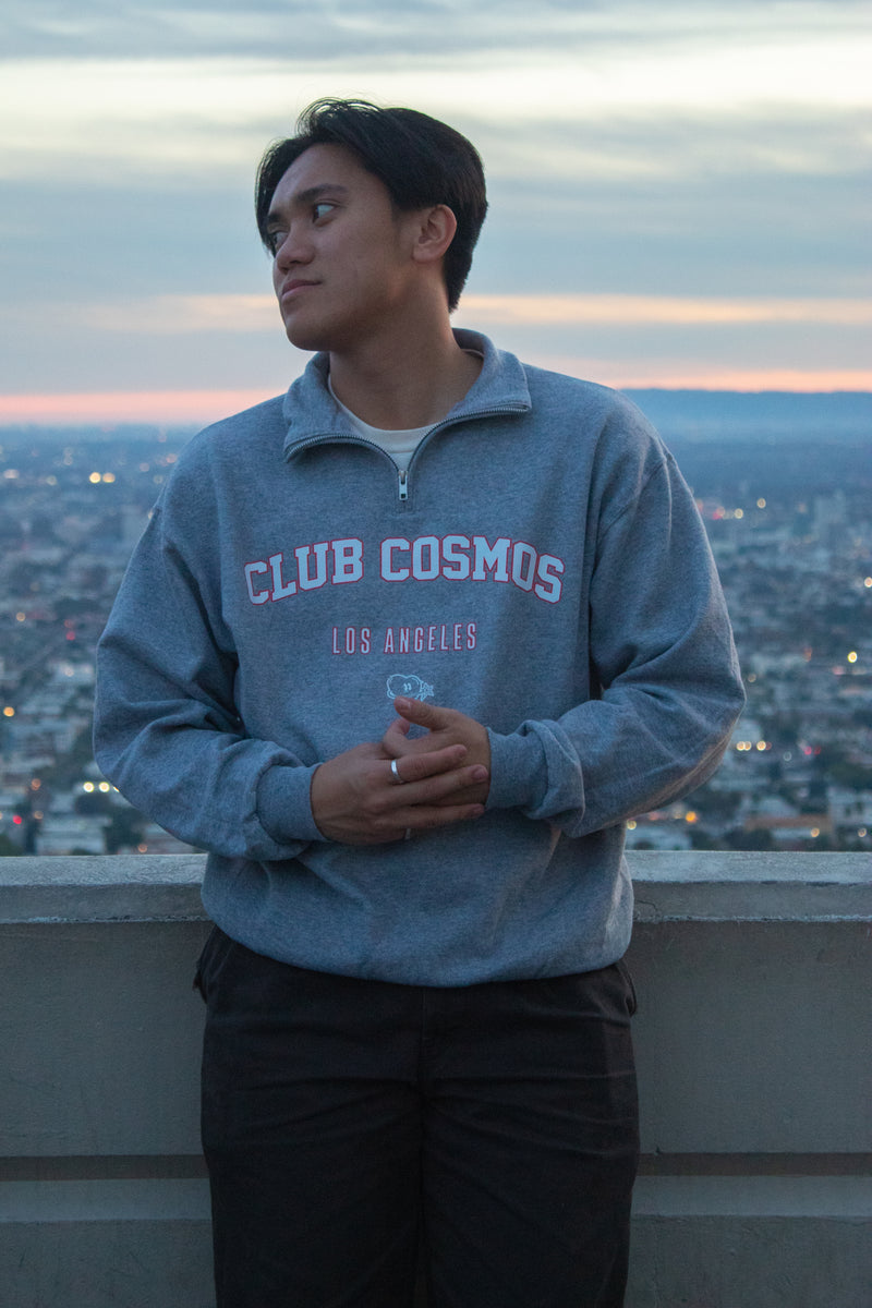 Model wearing a 'Club Cosmos Los Angeles' quarter zip sweatshirt with a cityscape in the background.