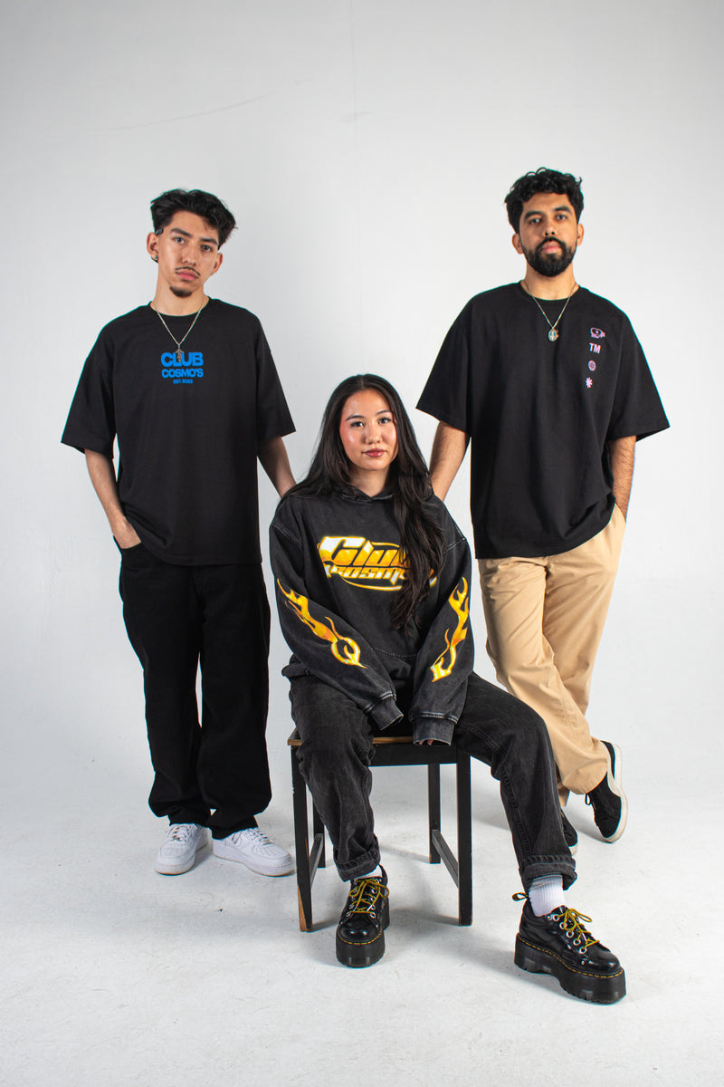 Three people total with two wearing black Club Cosmo's graphic tees with visible branding and one wearing a washed hoodie with orange and yellow Club Cosmo's graphics on a white background.