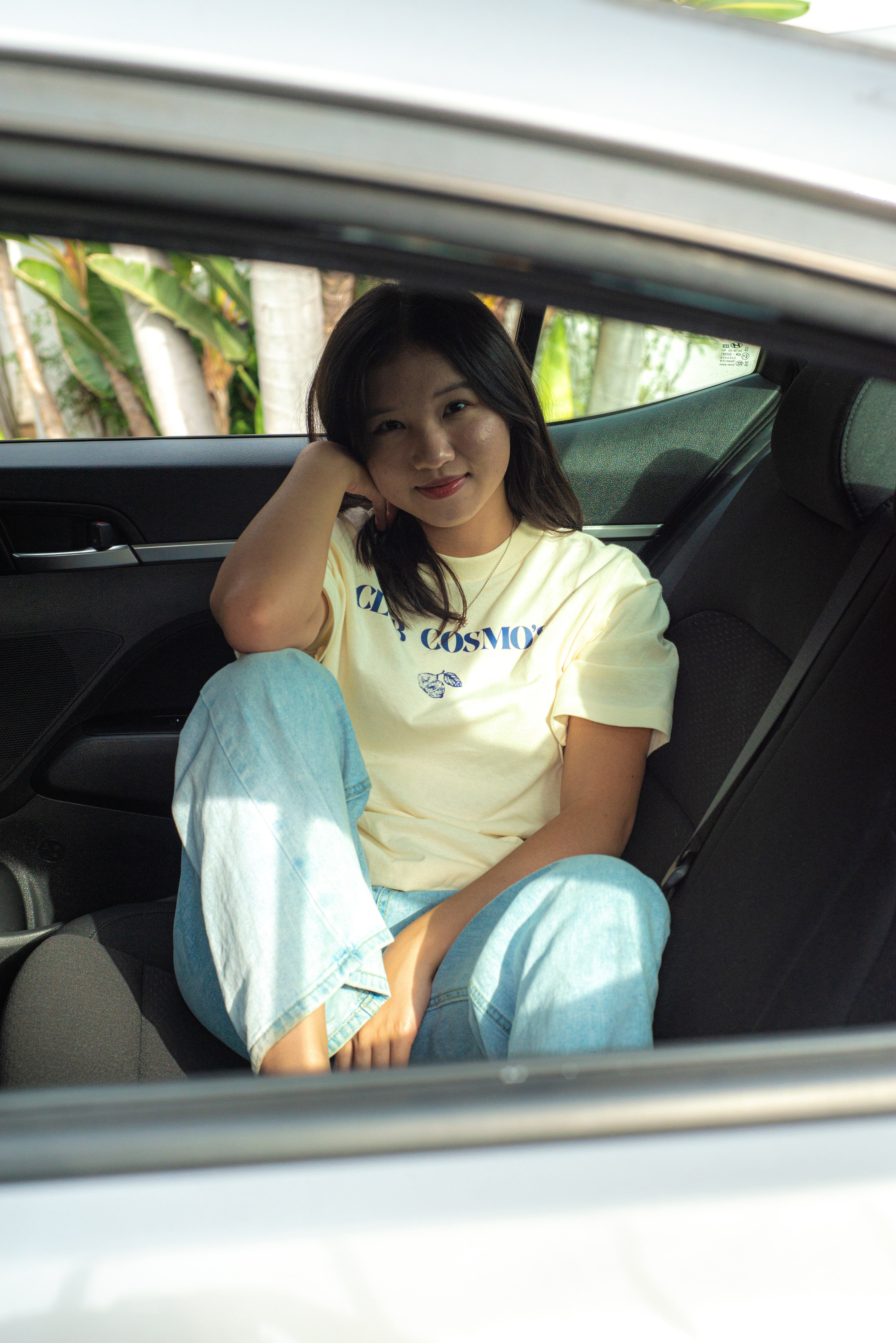 Model sitting in a car wearing a beige t-shirt with blue 'Club Cosmo's' text and light blue jeans.