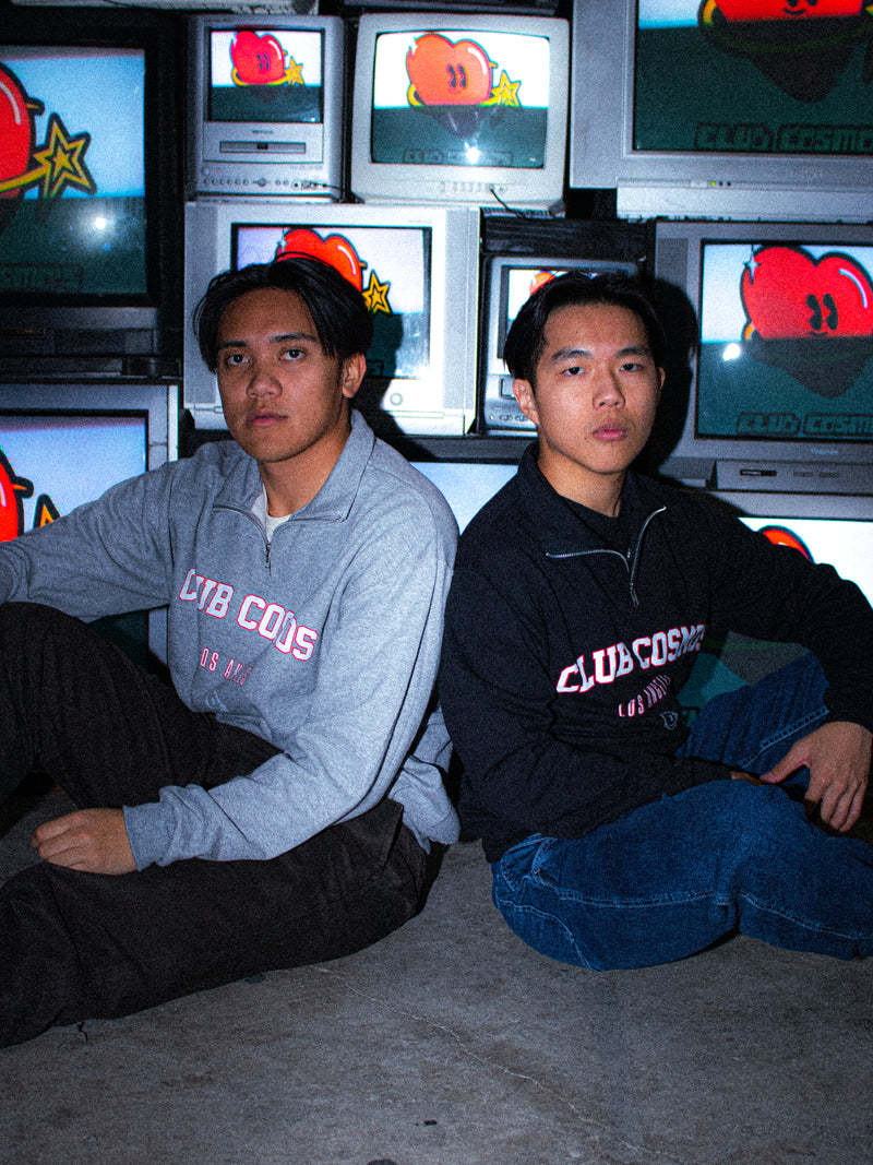Two individuals wearing a black and grey quarter zip sitting in front of multiple television monitors displaying the official Club Cosmo's logo.
