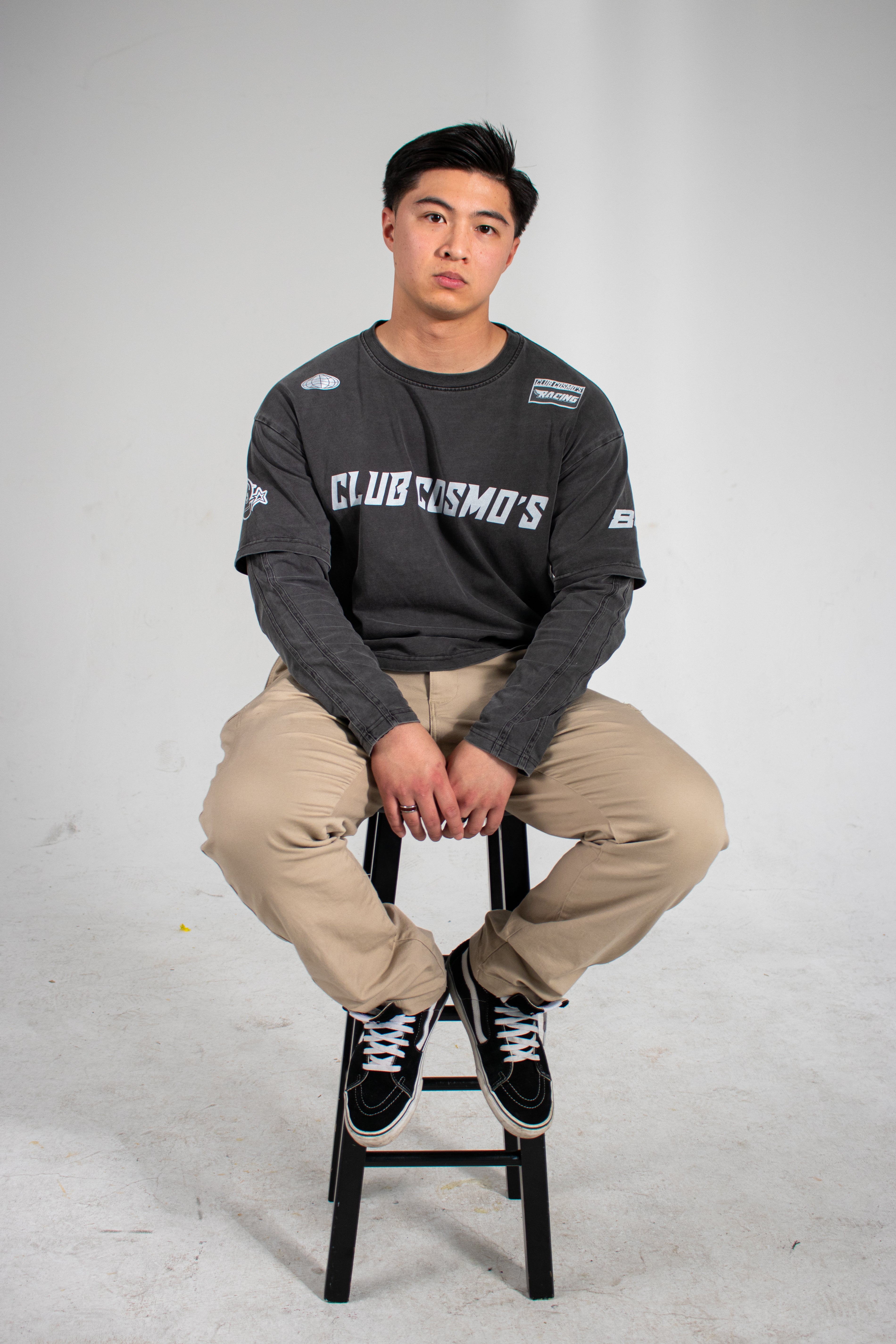 Model sitting on a stool wearing a dark long-sleeve shirt with Club Cosmo's branding and text with beige pants on a white background.