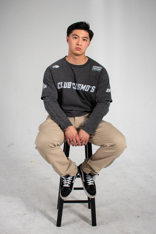 Model sitting on a stool wearing a dark long-sleeve shirt with Club Cosmo's branding and text with beige pants on a white background.