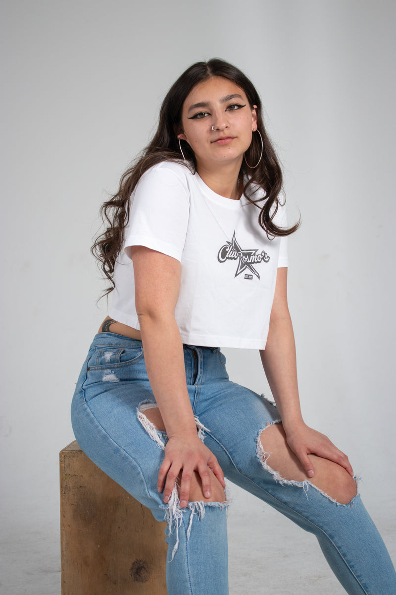 Model wearing a white t-shirt with a Club Cosmo's star logo and ripped blue jeans sitting on a wooden block against a plain white background.