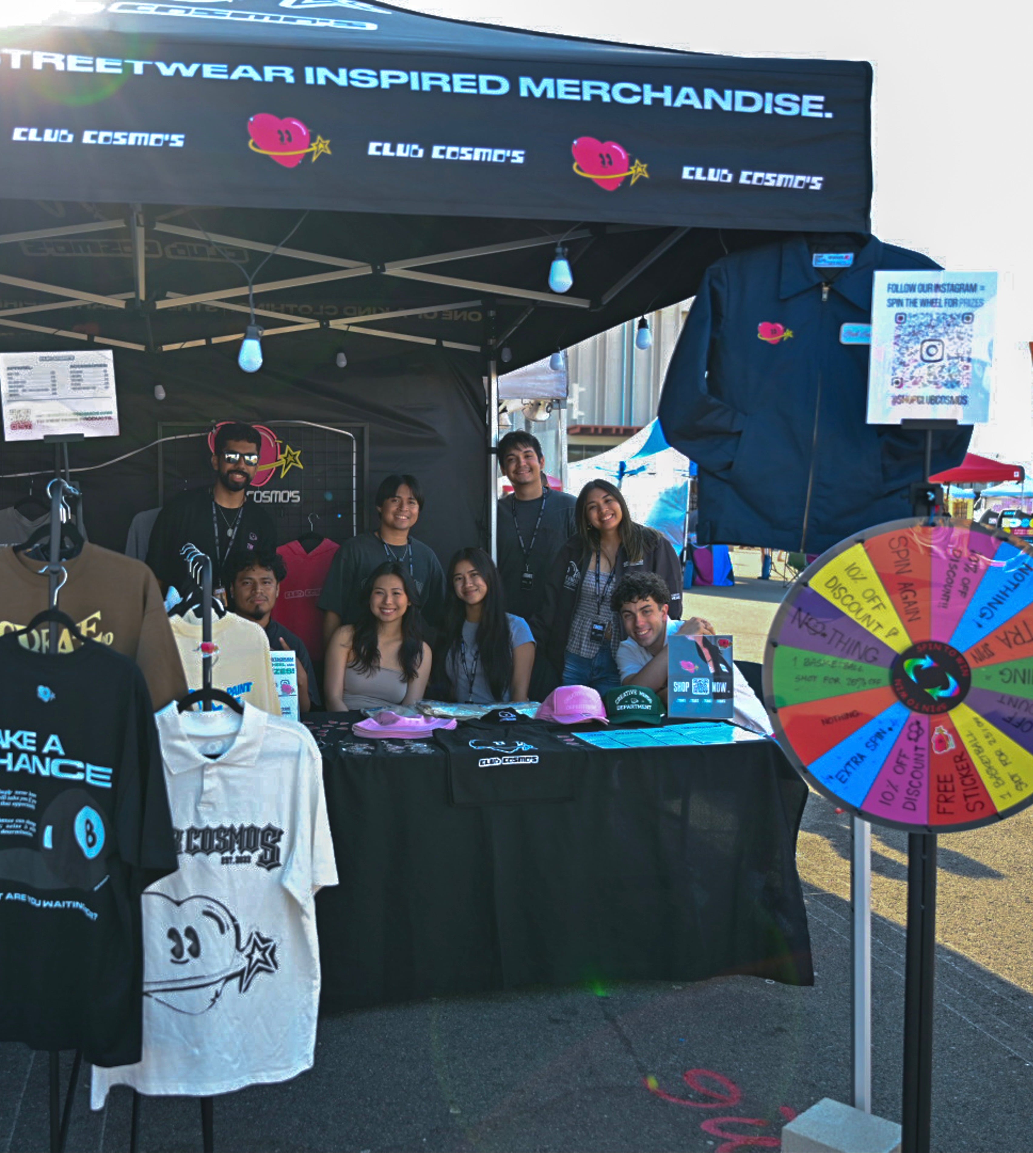 Outdoor night market event with a clothing booth displaying Club cosmo's merchandise and team members around.
