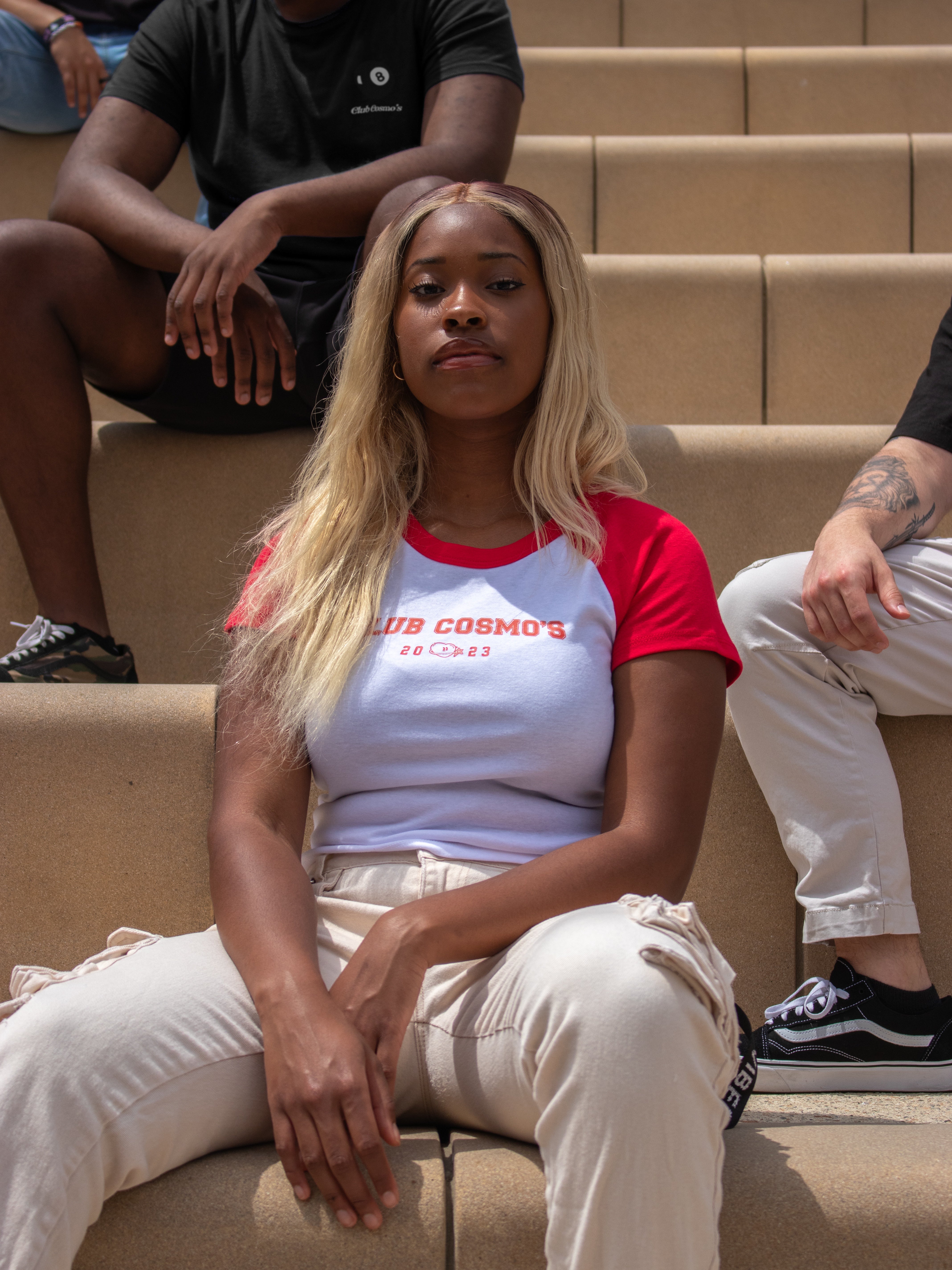 Female model wearing a red and white Club Cosmo's baby tee with text sitting on steps with additional models in the frame.
