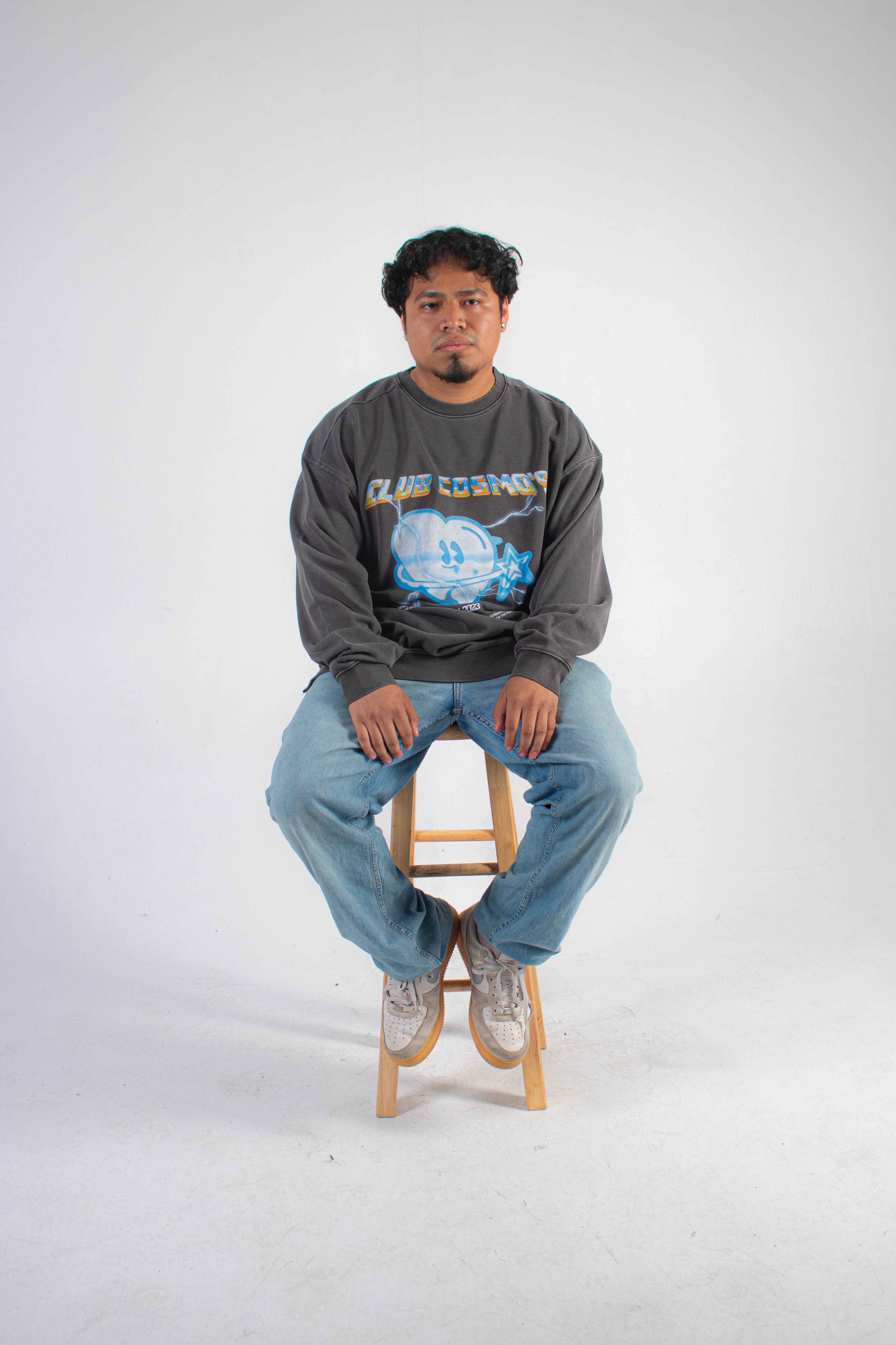 Model wearing a grey sweatshirt with a chrome Club Cosmo's logo graphic design sitting on a wooden stool against a white background.