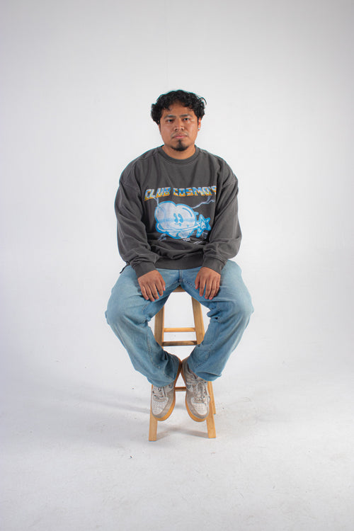 Model wearing a grey sweatshirt with a chrome Club Cosmo's logo graphic design sitting on a wooden stool against a white background.