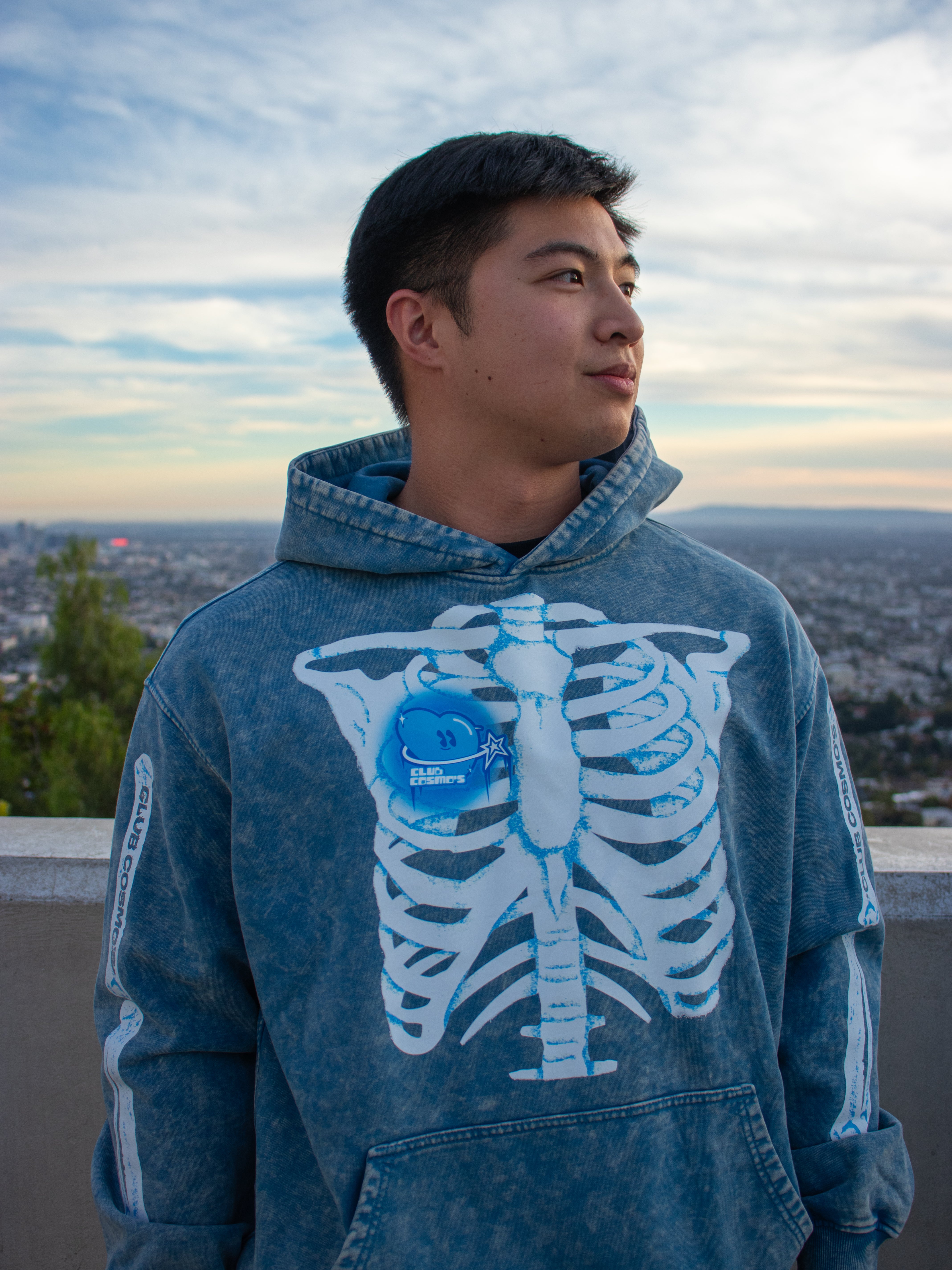 Model wearing a blue Club Cosmo's hoodie with a skeleton design outdoors.