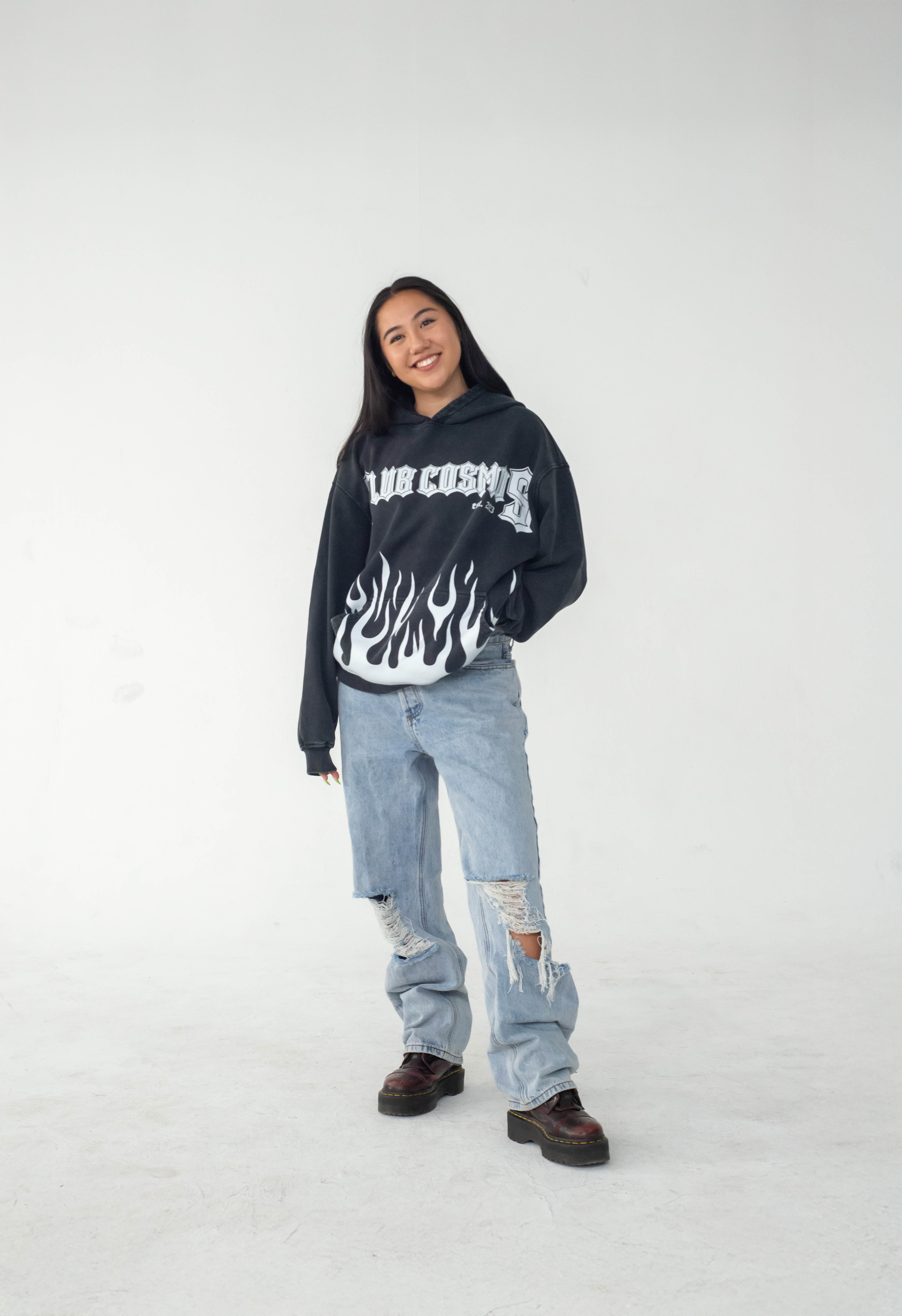 Model posing wearing a black hoodie with white text 'Club Cosmo's' and flames, light blue jeans, and brown red shoes on a white background.