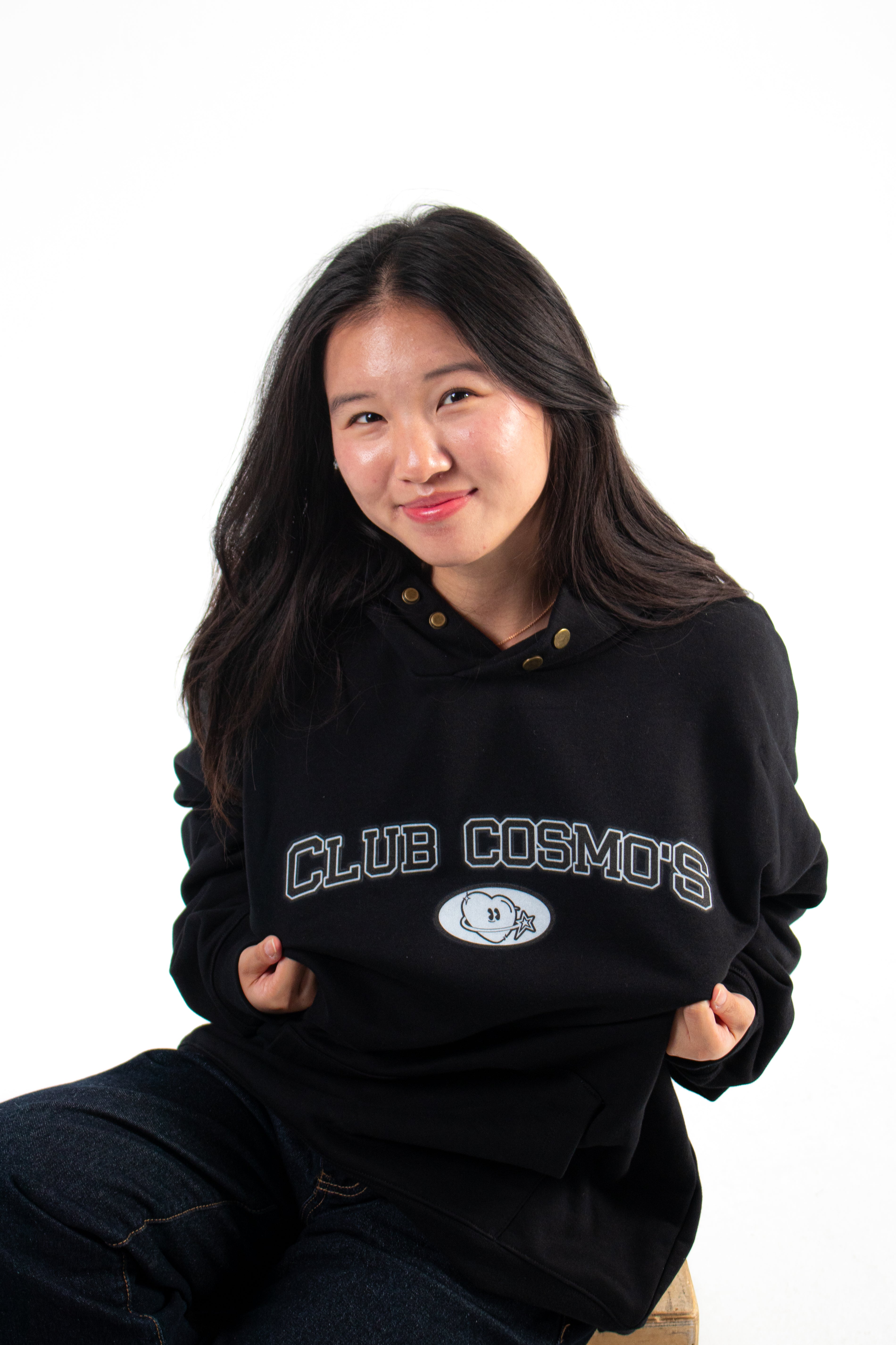 Model sitting down wearing a black hoodie with 'Club Cosmos' text on a white background.