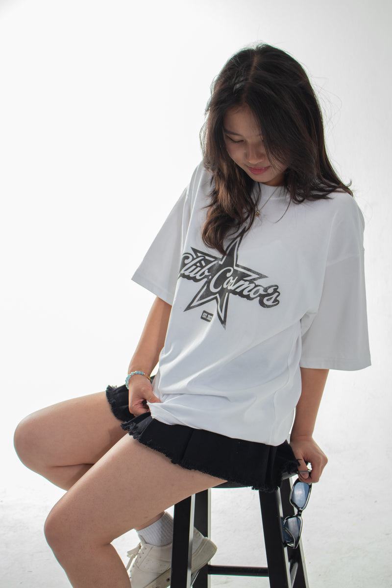 Model sitting on a stool wearing an oversized white t-shirt with a Club Cosmo's star logo on a white background.