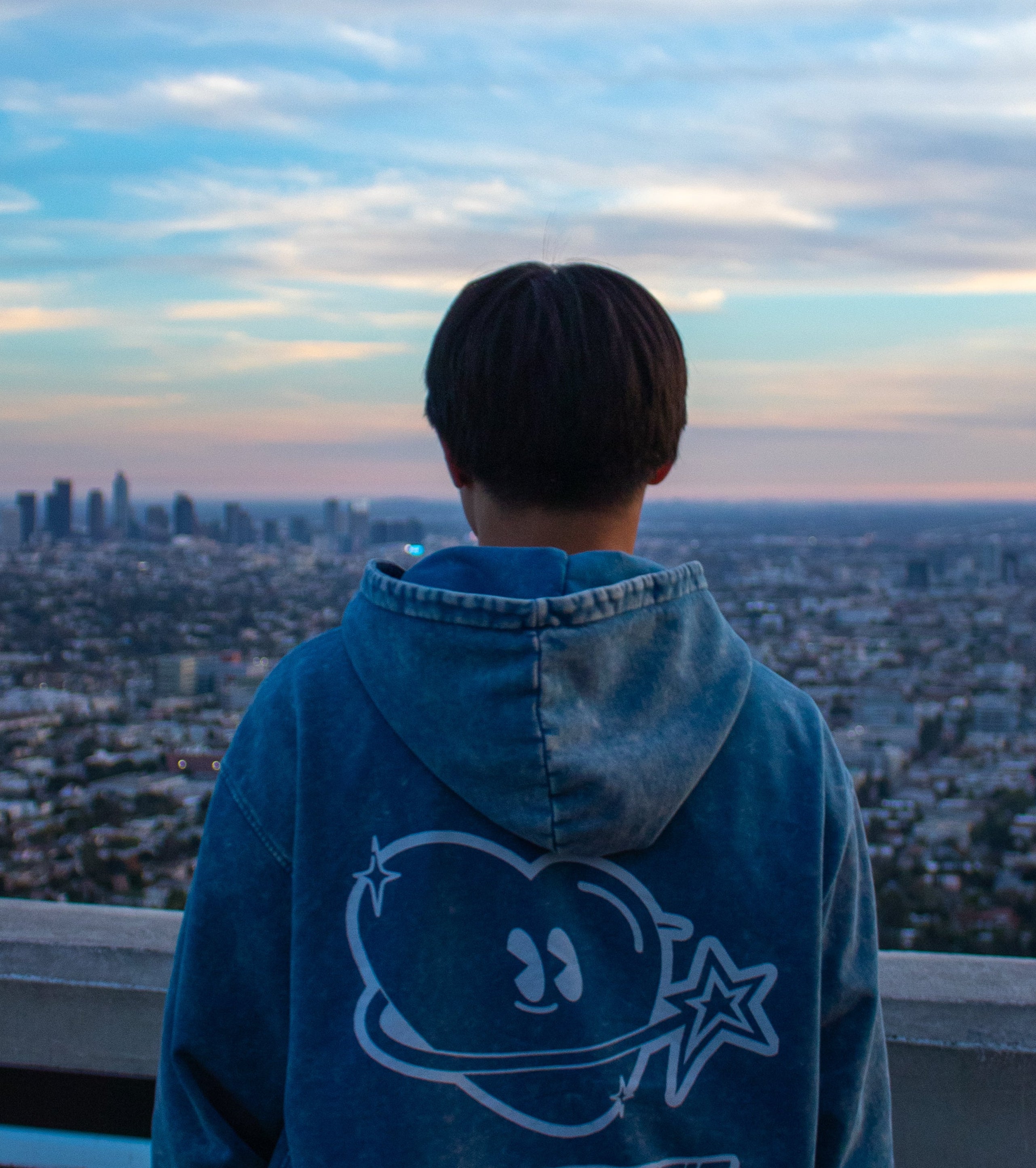 Person wearing a blue hoodie with 'Club Cosmo's' design, standing against a cityscape at sunset.