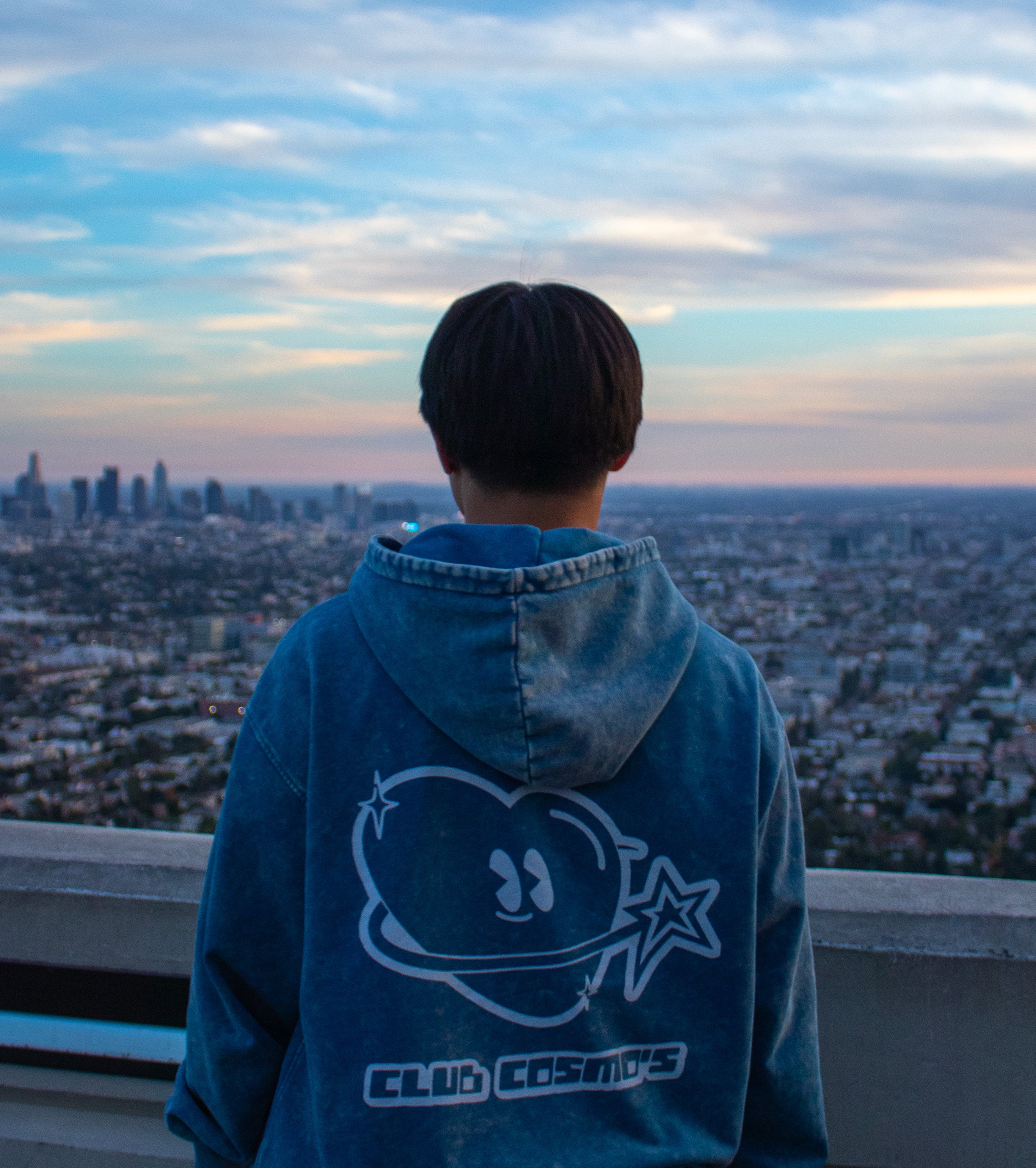 Person wearing a blue hoodie with 'Club Cosmo's' design, standing against a cityscape at sunset.