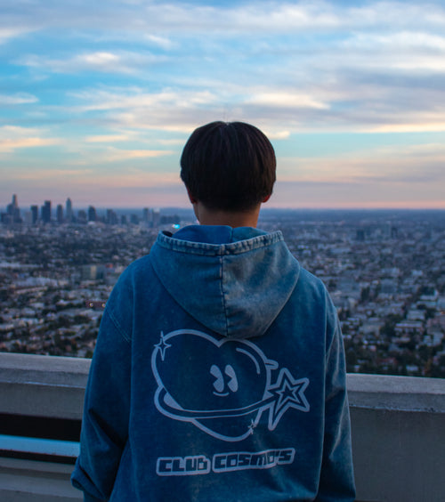 Person wearing a blue hoodie with 'Club Cosmo's' design, standing against a cityscape at sunset.